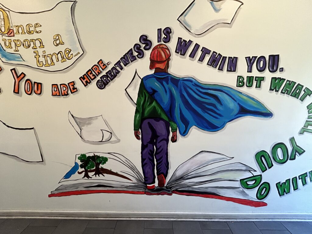 Boy with a blue cape and red hat, stand on an open book reading the purple words in a wall - Greatness is Within You.